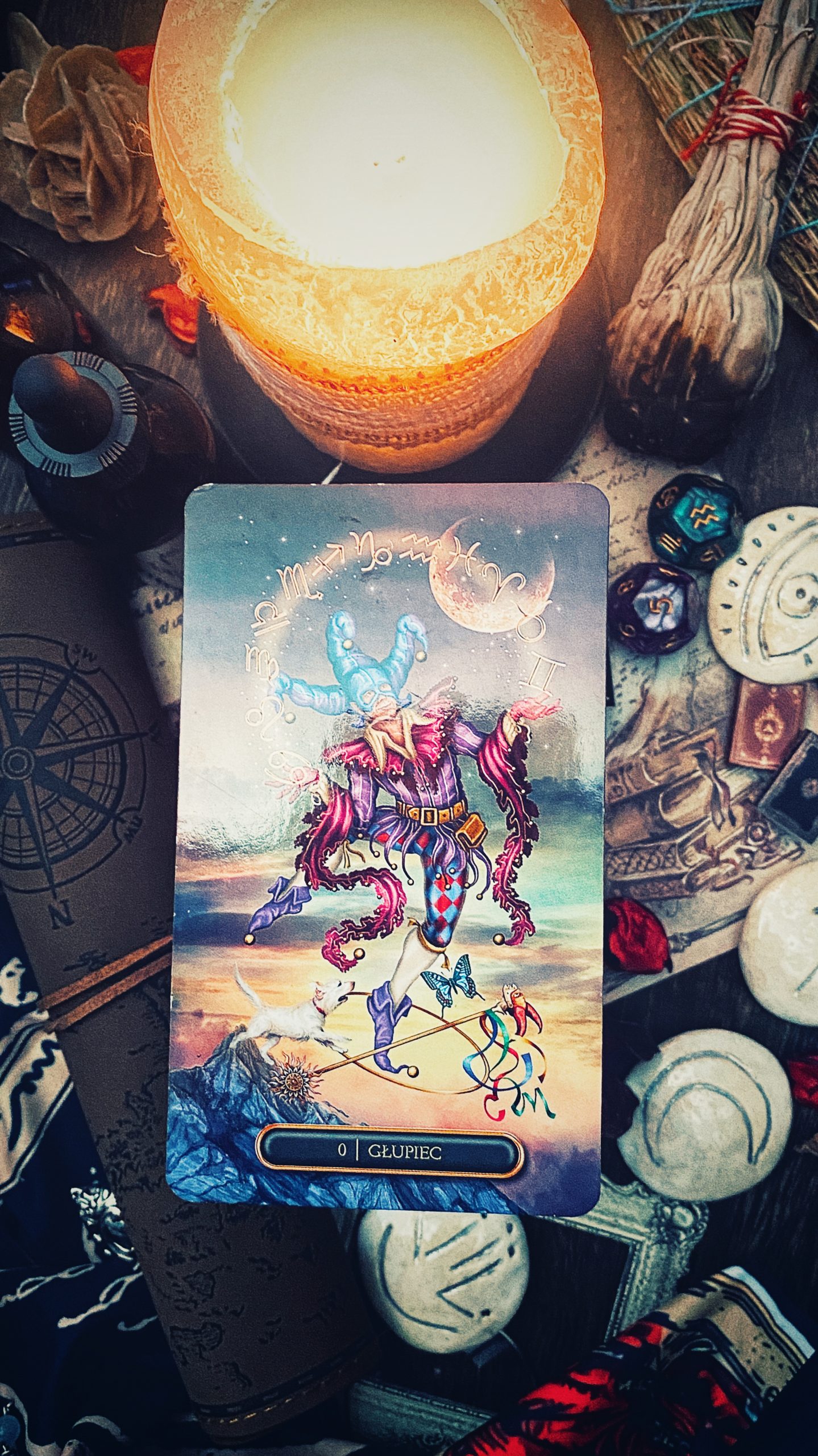 Tarot anielski postaw - karta Głupca w otoczeniu świecy i magicznych akcesoriów, symbolizująca nowe początki, niewinność i otwartość na nowe doświadczenia.